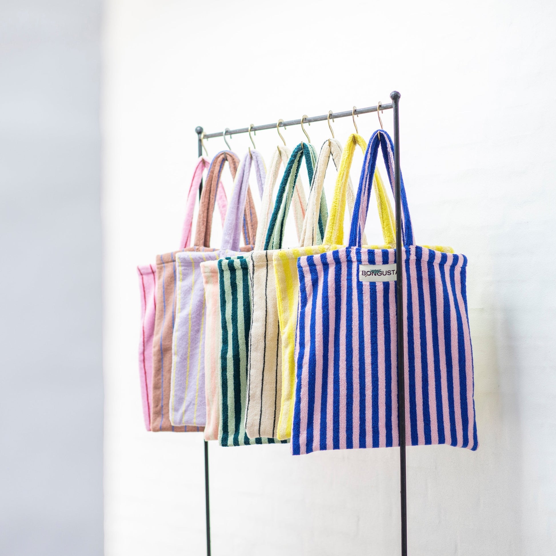 8 Bongusta classic tote bags hang on rustic gold hangers on a black rail. The bags are pink and thin neon striped, brown and thin blue striped, lilac and thin neon yellow striped, cream and pink striped, light and dark green striped, cream and thin black striped, cream and neon yellow striped and a pink and neon blue striped bag. 
