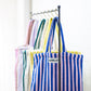 8 Bongusta classic tote bags hang on rustic gold hangers on a black rail. The bags are pink and thin neon striped, brown and thin blue striped, lilac and thin neon yellow striped, cream and pink striped, light and dark green striped, cream and thin black striped, cream and neon yellow striped and a pink and neon blue striped bag. 
