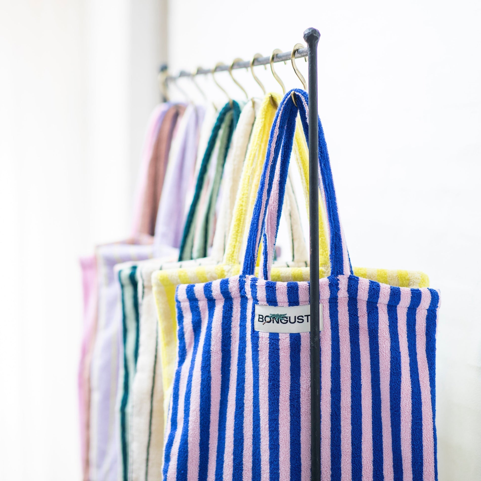8 Bongusta classic tote bags hang on rustic gold hangers on a black rail. The bags are pink and thin neon striped, brown and thin blue striped, lilac and thin neon yellow striped, cream and pink striped, light and dark green striped, cream and thin black striped, cream and neon yellow striped and a pink and neon blue striped bag. 