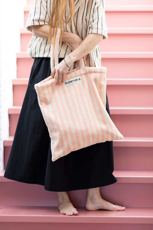 A cream and pink striped classic tote bag, made of towelling fabric with a long pink and cream handle being held by someone standing on pink steps without shoes on wearing a long black skirt and a cream and thin striped black button up shirt made of towelling fabric. At the top of the bag it says BONGUSTA in black inside of a cream box. 