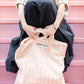 A cream and pink striped classic tote bag, made of towelling fabric with a long pink and cream handle being held by someone sitting on pink steps wearing a long black skirt and a cream and thin striped black button up shirt made of towelling fabric. At the top of the bag it says BONGUSTA in black inside of a cream box. 