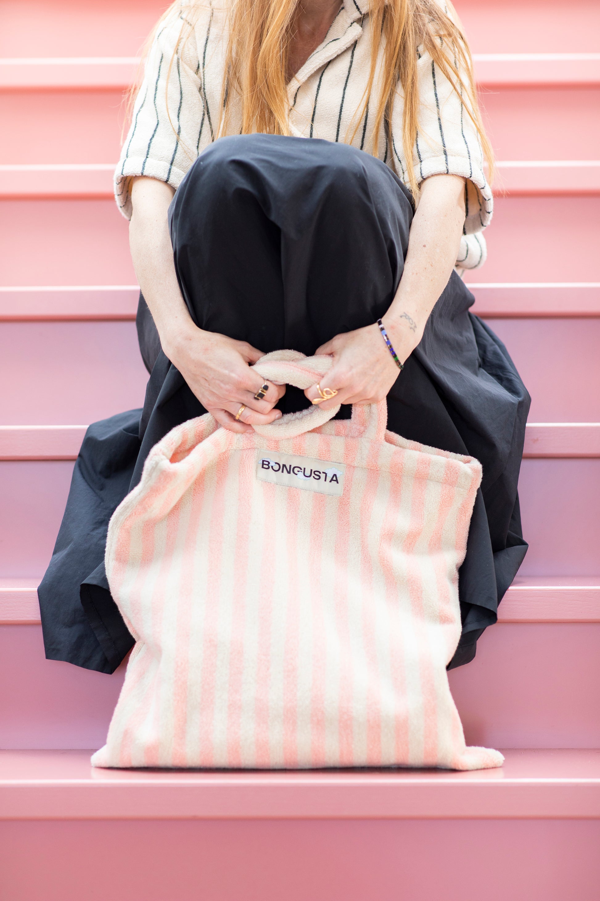 A cream and pink striped classic tote bag, made of towelling fabric with a long pink and cream handle being held by someone sitting on pink steps wearing a long black skirt and a cream and thin striped black button up shirt made of towelling fabric. At the top of the bag it says BONGUSTA in black inside of a cream box. 