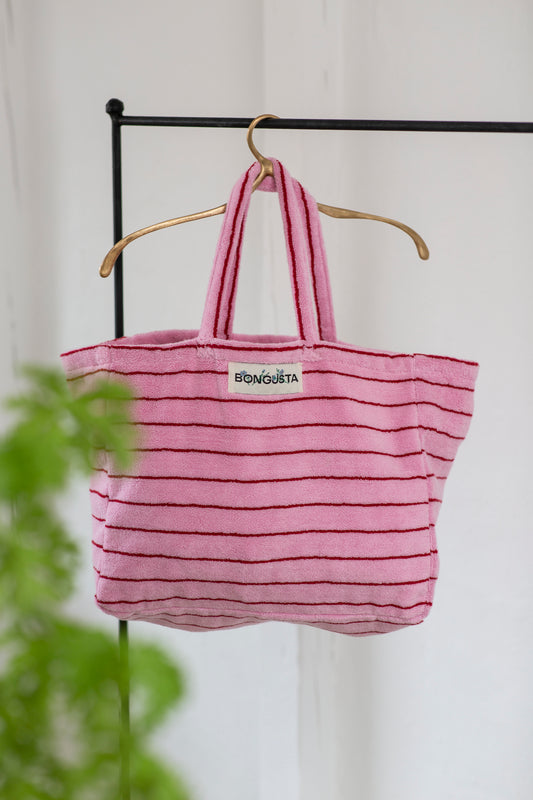 A light pink weekend style tote bag with thin red stripes, made of towelling fabric with a long pink and red striped handle hangs on a rusty gold hanger on a black rail. At the top of the bag it says BONGUSTA in black with green flowers coming off the letters, inside of a cream box. 