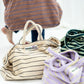 A cream weekend style tote bag with thin black stripes, made of towelling fabric with a long cream and black striped handle sits on white flooring next to the same bags, one lilac one with thin yellow neon stripes and one bag that has dark and light green stripes on it. Above these bags, being held by someone wit dark blue jeans and no shoes on is a brown bag with thin blue neon stripes. At the top of the bag it says BONGUSTA in black with green flowers coming off the letters, inside of a cream box. 