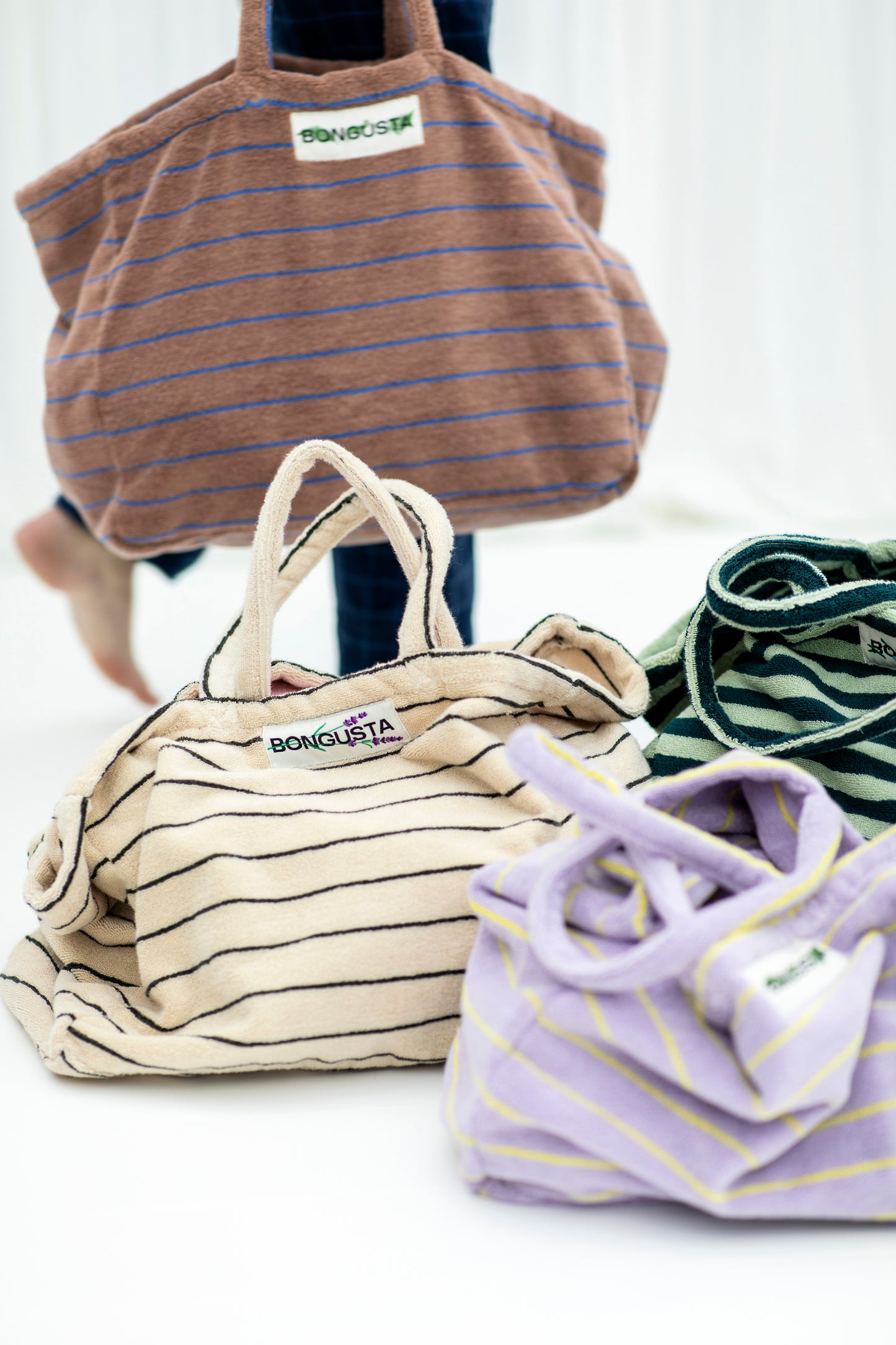 A cream weekend style tote bag with thin black stripes, made of towelling fabric with a long cream and black striped handle sits on white flooring next to the same bags, one lilac one with thin yellow neon stripes and one bag that has dark and light green stripes on it. Above these bags, being held by someone wit dark blue jeans and no shoes on is a brown bag with thin blue neon stripes. At the top of the bag it says BONGUSTA in black with green flowers coming off the letters, inside of a cream box. 