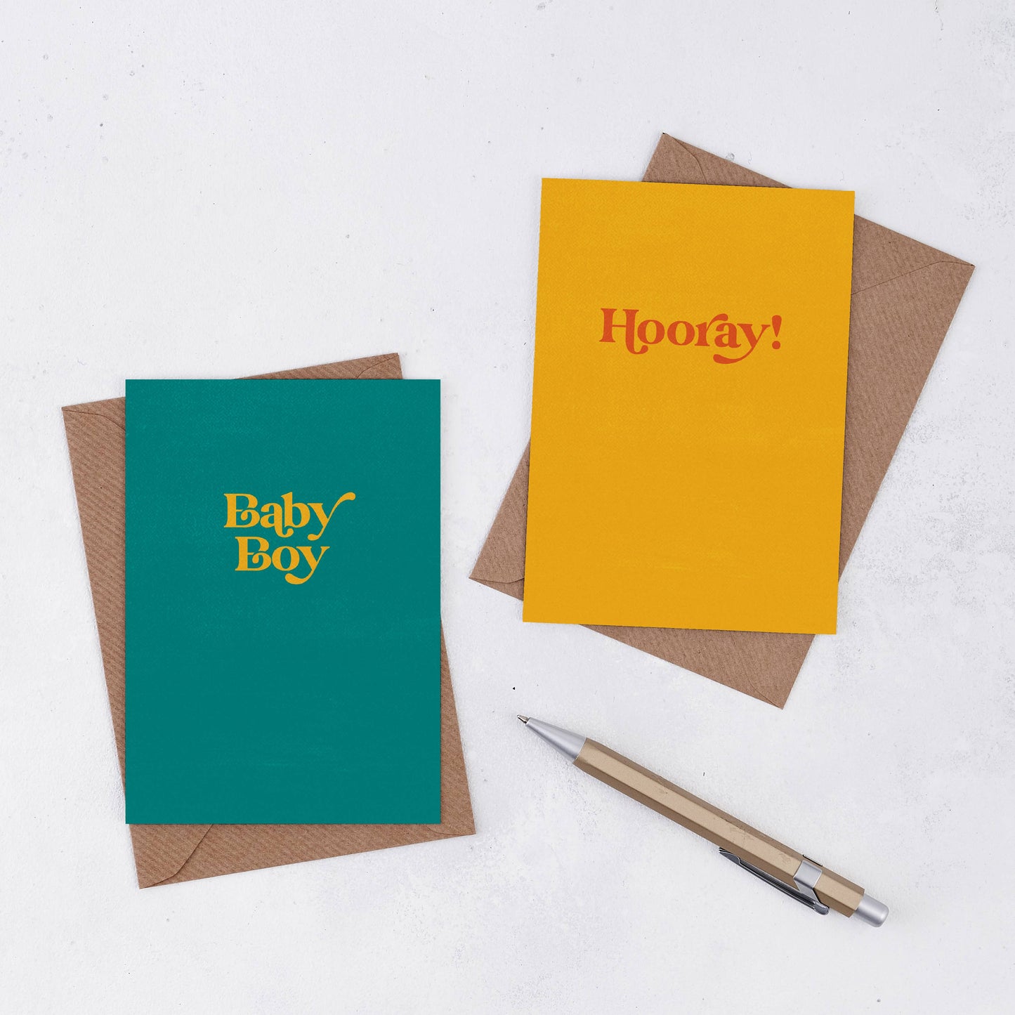 Two simple greeting cards on brown Kraft envelopes. One is teal with eh words Baby Boy in yellow, and one is yellow with the words Hooray! in red in a retro font