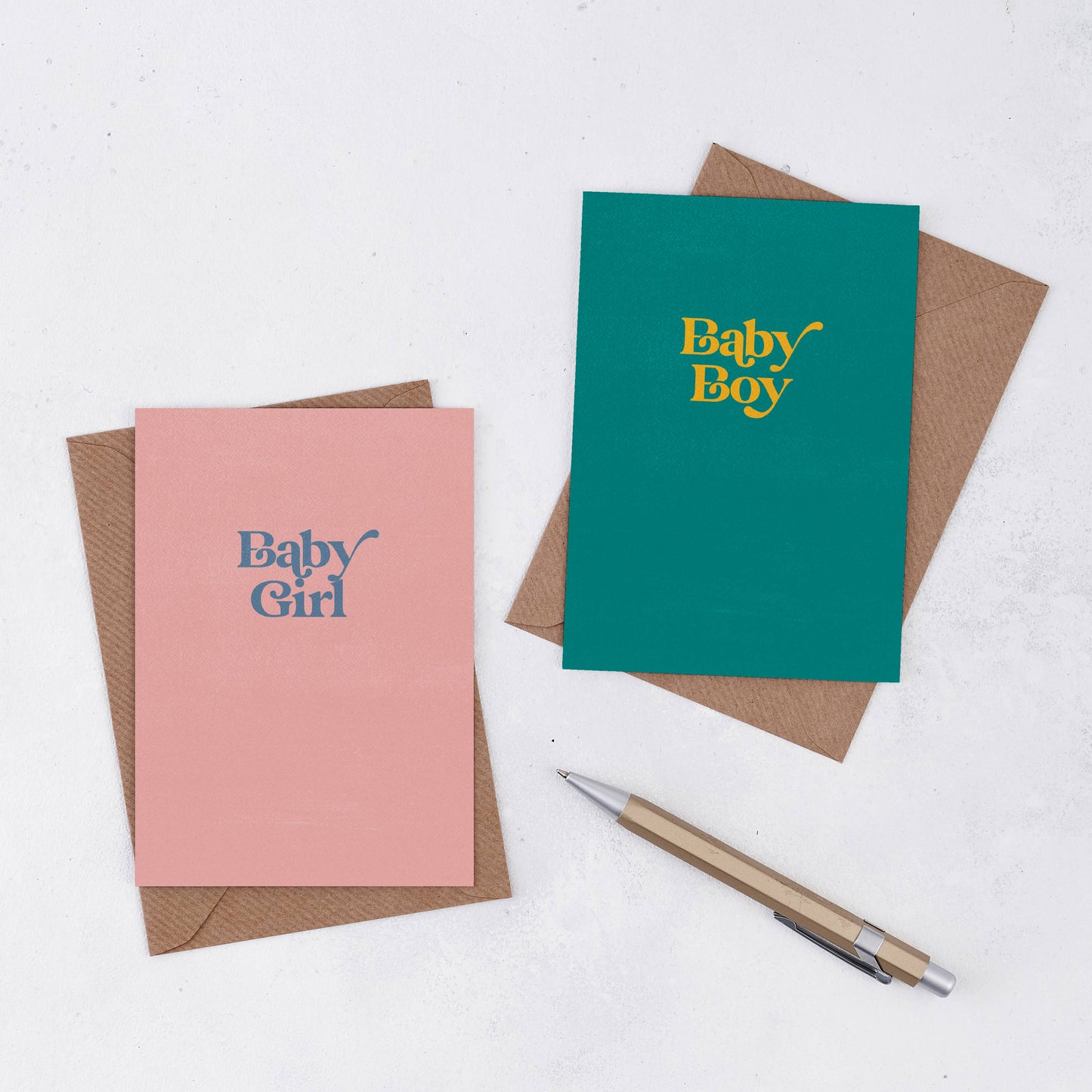 Two simple greetings cards next to brown Kraft envelopes. One is teal with the words Baby Boy in yellow, and one is pink with the words Baby Girl in grey in a retro font