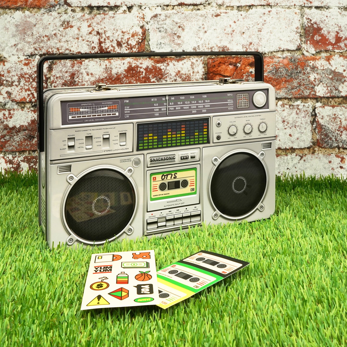 Boombox Lunch Box – retro ghetto blaster-inspired tin lunch box with an animated lenticular graphic equalizer, embossed details, and a realistic carry handle. Includes stickers for personalisation. Perfect for carrying snacks and lunches in style, suitable for both kids and adults. Shown sat against a brick wall onsite grass with the customisable stickers