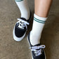 Black sneakers with white laces worn with white socks featuring green stripes on a concrete floor.