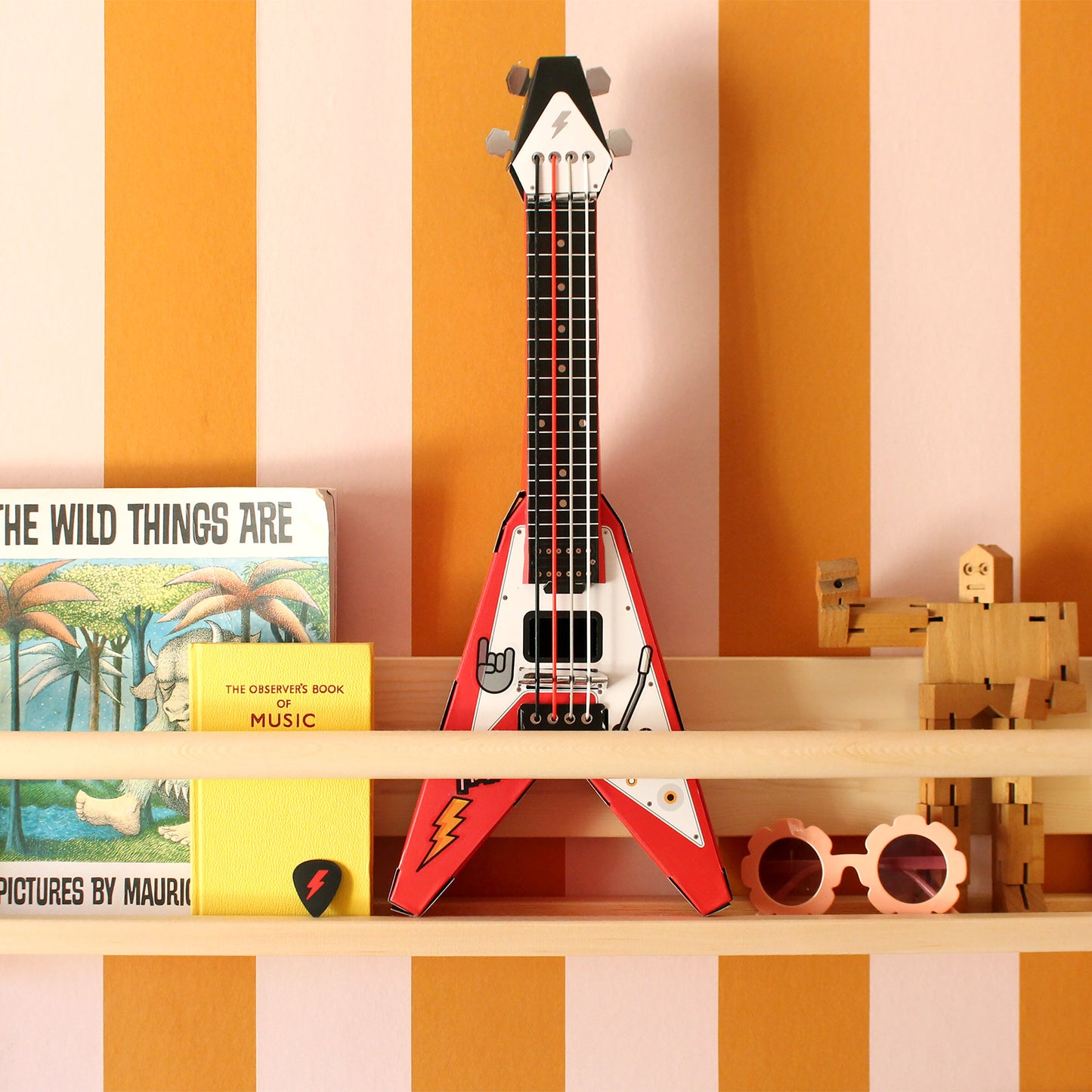 Red Build Your Own Rock Star Guitar – a playable cardboard Flying-V guitar kit featuring pop-out components that assemble without glue or tape. Elastic bands serve as strings, making it a fun DIY project for kids to create their own unique musical instrument. Sits on a wooden bookshelf against mustard and pink striped wallpaper