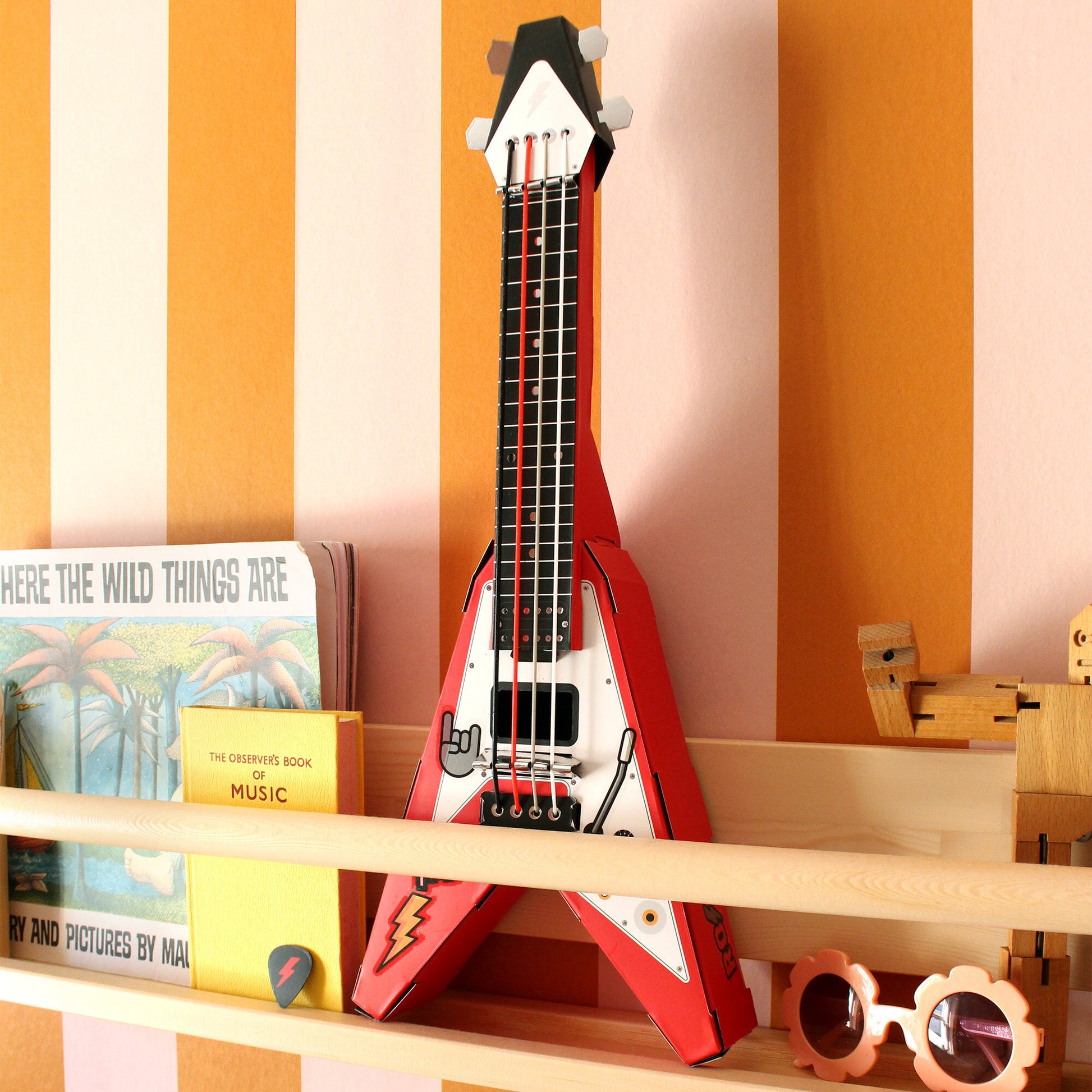 Red Build Your Own Rock Star Guitar – a playable cardboard Flying-V guitar kit featuring pop-out components that assemble without glue or tape. Elastic bands serve as strings, making it a fun DIY project for kids to create their own unique musical instrument. Sits on a wooden bookshelf against mustard and pink striped wallpaper