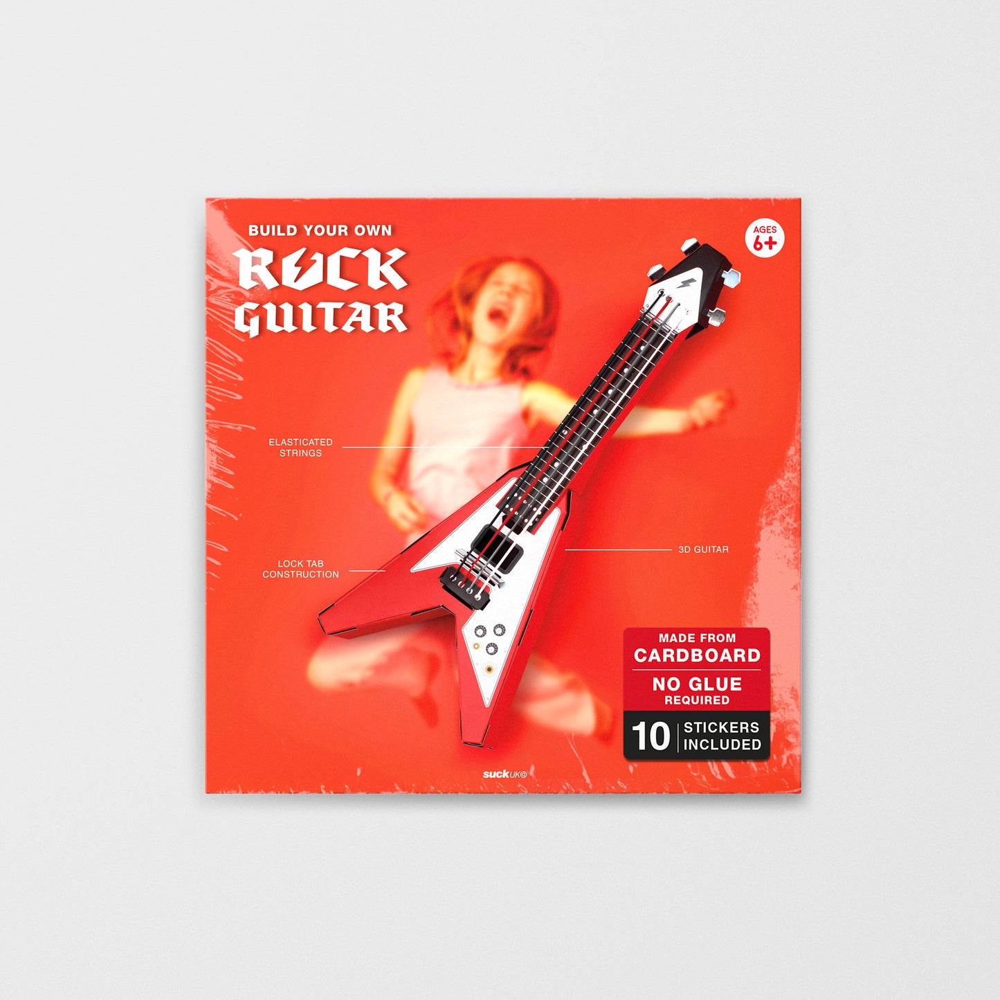 Red Build Your Own Rock Star Guitar – a playable cardboard Flying-V guitar kit featuring pop-out components that assemble without glue or tape. Elastic bands serve as strings, making it a fun DIY project for kids to create their own unique musical instrument. Sits on a wooden bookshelf against mustard and pink striped wallpaper. Shows the red packet which the kit comes in