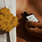 Hand holding a perfume bottle labeled 'cruissant cafe' next to a yellow floral arrangement.