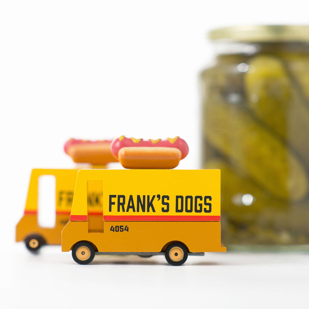 Toy hot dog truck labeled 'Frank's Dogs' next to a jar of pickles on a white background