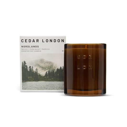 Cedar London Nordlands Candle in a refillable amber glass jar, embossed with 'CDR LDN,' next to its packaging featuring a Nordic landscape with a forest and mountain lake. Scent notes include spruce, cedarwood, oak moss, pine, and eucalyptus.