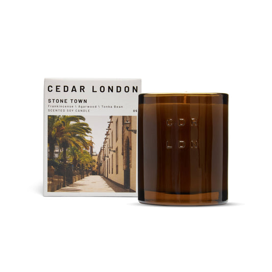 Cedar London Stone Town Candle in a refillable amber glass jar, embossed with 'CDR LDN,' next to its packaging featuring a sunlit, historic street scene. Scent notes include frankincense, agarwood, tonka bean, vanilla, and patchouli.