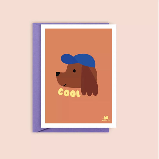 A greeting card with a warm terracotta background featuring an illustrated brown dog wearing a blue cap. The word "COOL" is printed in playful, yellow vintage-style text below the dog's face. The card is paired with a pastel purple envelope.