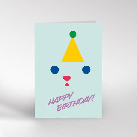 An A6 greeting card with a light blue background. Against it is two blue eyes and a pink nose with a yellow party hat above the eyes creating a face with happy birthday written underneath the face in purple writing. 