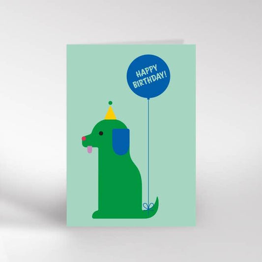 An A6 greeting card with the image of a green dog sitting down with a yellow party hat on and a blue balloon attached to its tail saying happy birthday. 