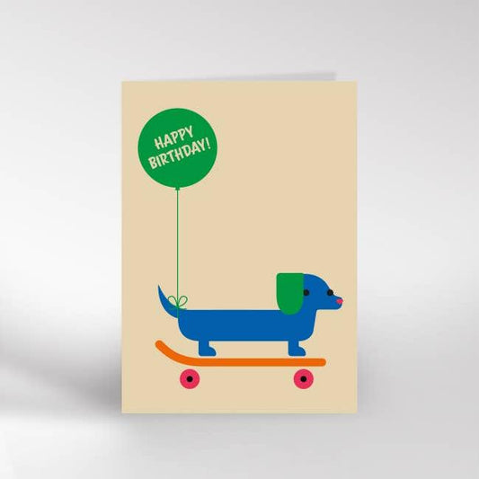 An A6 greeting card with a blue sausage dog on an orange skateboard with a green balloon tied to its tail that says happy birthday 