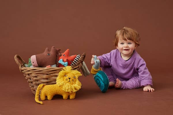 On a rust coloured backdrop a toddler in a purple top and bottoms is playing with the knitted Donna Wilson toys out of a woven basket. In the basket is a horse and fox toy, in front is a lion and the child is holding the snail