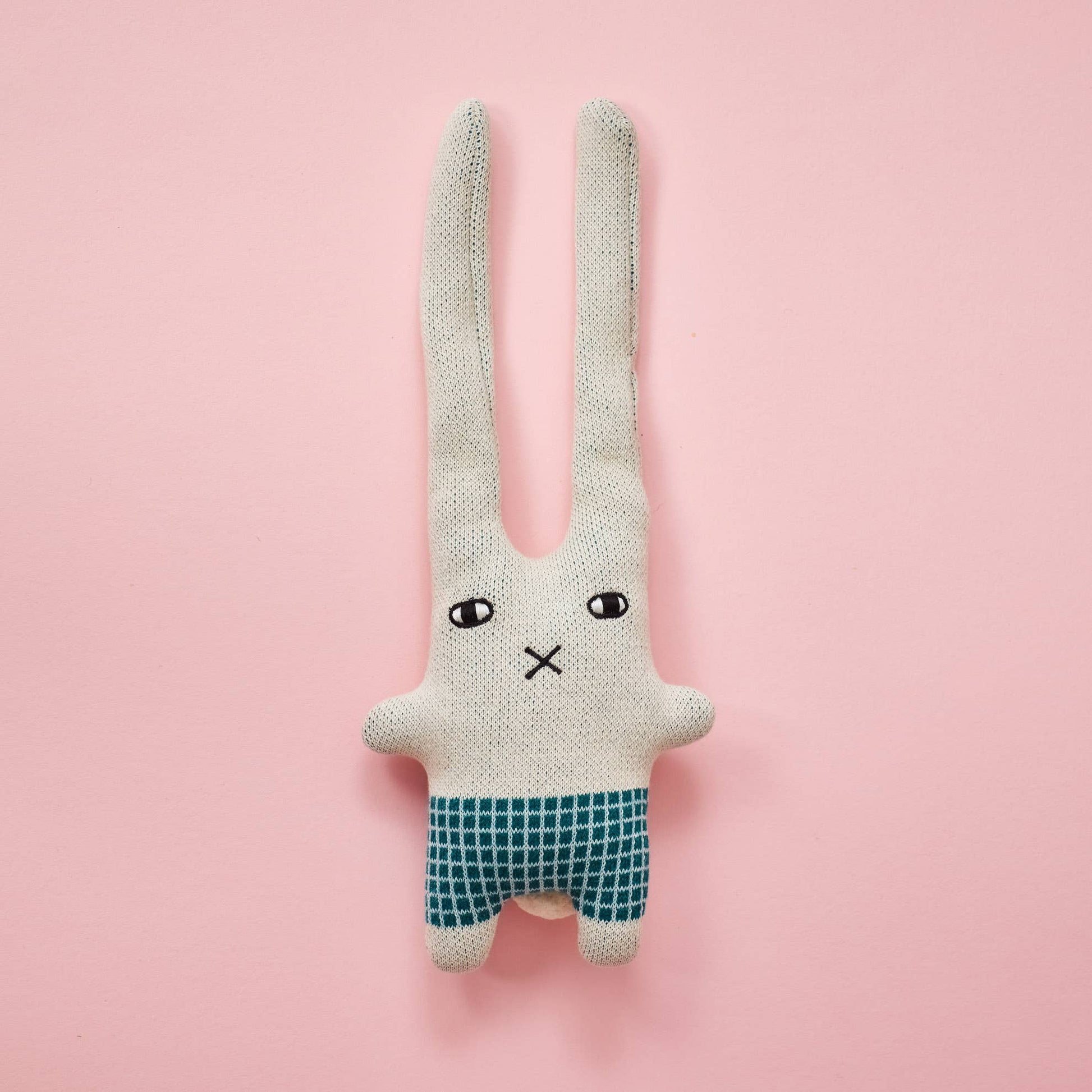 Donna Wilson knitted wool bunny in a naive illustration style. It has a square body with green check shorts and short little arms and feet, with two extra long ears. A little embroidered cross nose and white and black eyes, on a light grey base. And a pink with green lettering label spelling Donna Wilson, all photographed on a pink backdrop