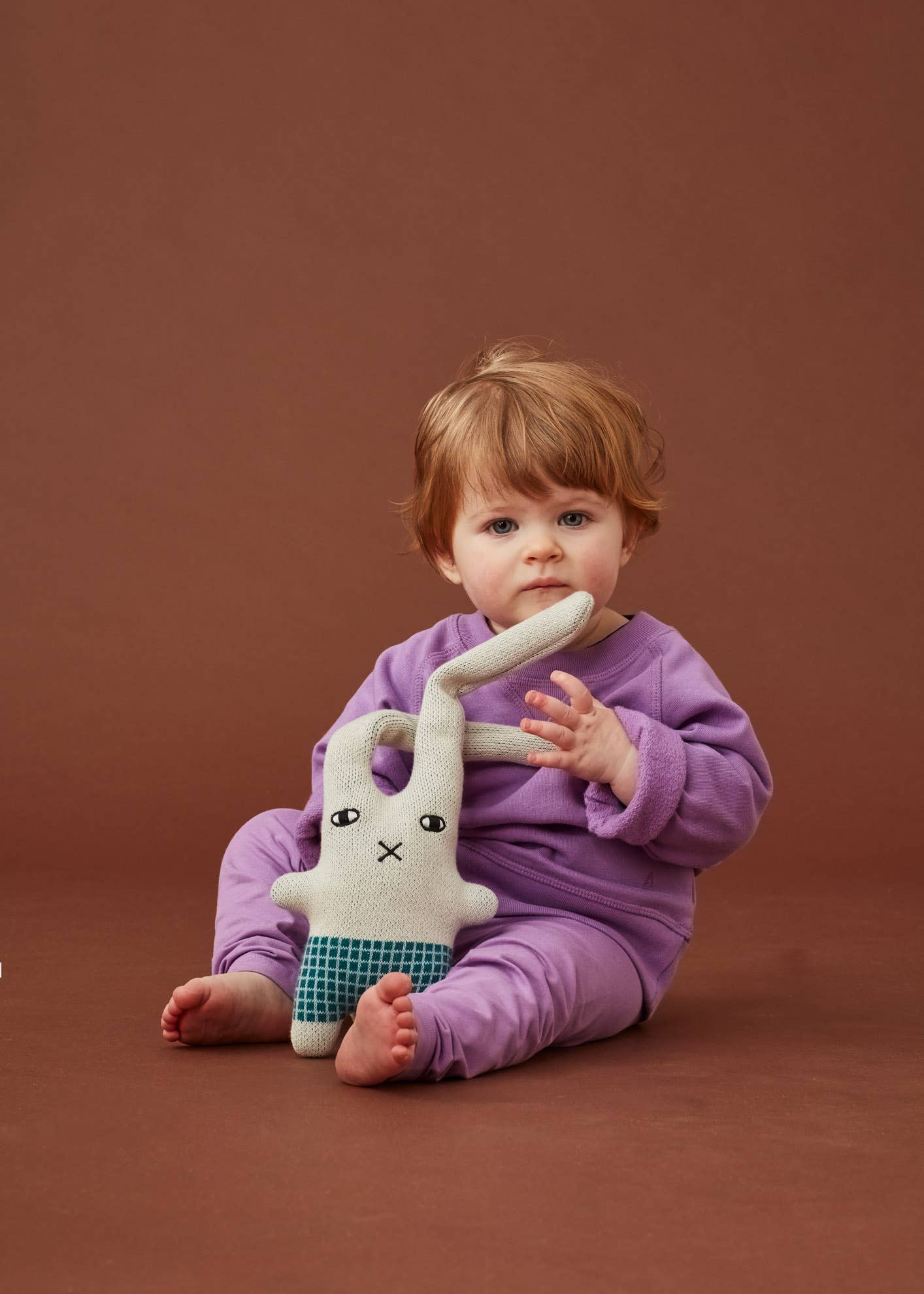 A toddler in a Purple matching top and trousers, on a rust colour backdrop holding the Donna Wilson knitted wool bunny in a naive illustration style. It has a square body with green check shorts and short little arms and feet, with two extra long ears. A little embroidered cross nose and white and black eyes, on a light grey base. And a pink with green lettering label spelling Donna Wilson