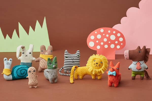 On a rust backdrop with paper cutout grass, clouds and toast stall in the background. All of the Donna Wilson small knitted toys, from left to right is a snail,  bunny, peanut, mole, raccoon, cat, lion, fox and horse, all in their naive illustrated style