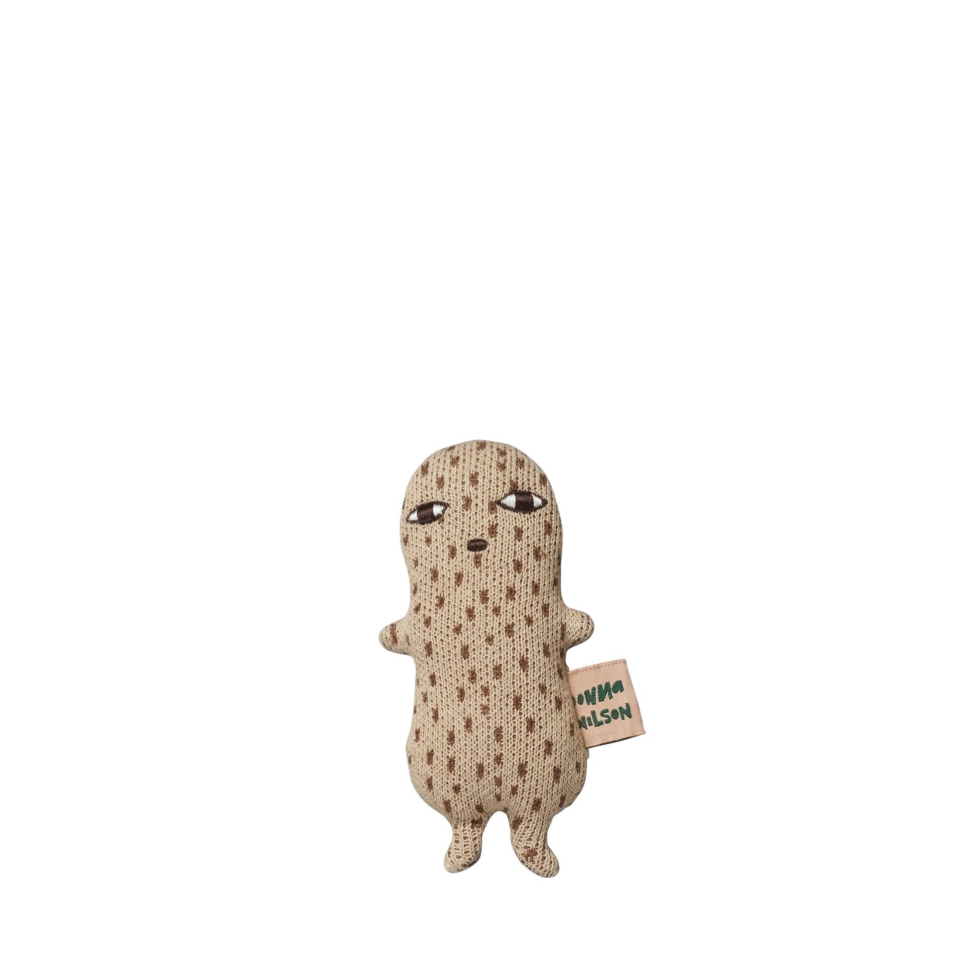 Knitted peanut rattle by Donna Wilson in a naive illustrated style, in a creamy light brown colour with brown dash lines all over and a little embroidered mouth and eyes. On the right side is a small pink tag with green lettering spelling Donna Wilson 