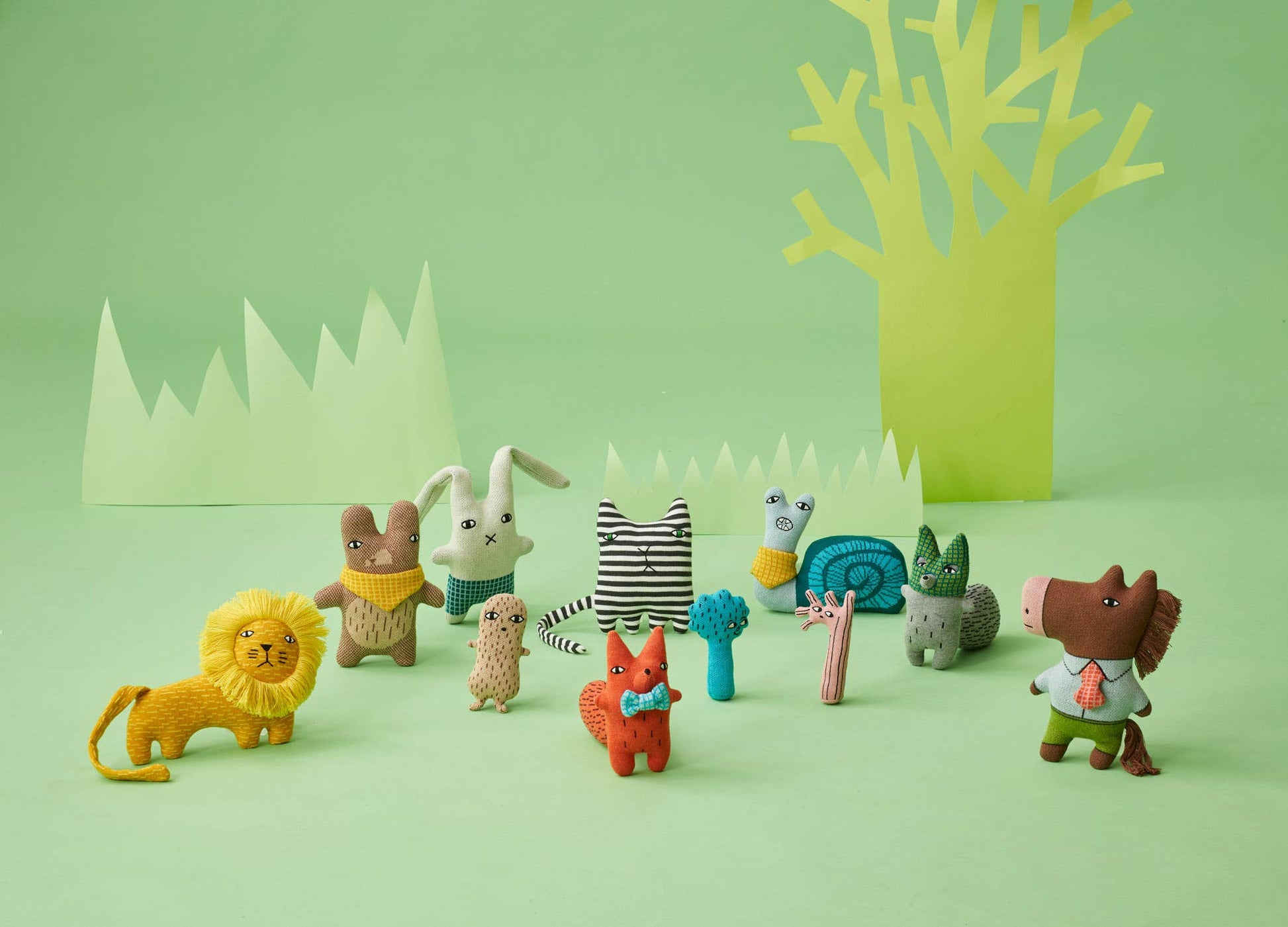 On a bright green backdrop with grass and tree paper cutouts, from left to right is all of the Donna Wilson knitted toys. A lion, mole, bunny, peanut, fox, cat, trees, snail. Raccoon, and horse