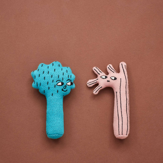 Two little mini greet rattles in the naive illustration style by Donna Wilson. One on the left is pink with bark lines and it has five small branches off of its head next to two little eyes and a mouth. On the right is a blue tree with bark lines, with a spikes round head and two little eyes and smiley mouth. They both have a pink tag on the right side with green text spelling Donna Wilson. All on a rust backdrop