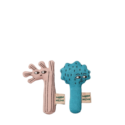 Two little mini greet rattles in the naive illustration style by Donna Wilson. One on the left is pink with bark lines and it has five small branches off of its head next to two little eyes and a mouth. On the right is a blue tree with bark lines, with a spikes round head and two little eyes and smiley mouth. They both have a pink tag on the right side with green text spelling Donna Wilson