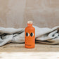 Orange bottle with black eye design on a wooden surface with a beige background