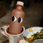 Brown bottle with face design on a table with plates and a cup.
