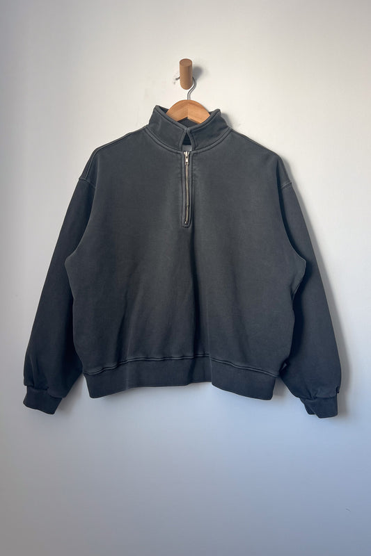 A charcoal-coloured sweatshirt with a quarter-zip design, featuring a high collar and long sleeves. The sweatshirt has a relaxed fit and ribbed cuffs and hem, displayed on a wooden hanger against a plain light grey wall.