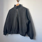 A charcoal-coloured quarter-zip sweatshirt hanging on a wooden hanger against a light grey wall. The sweatshirt has a relaxed fit, high collar, long sleeves, and ribbed cuffs and hem, with a visible side view.