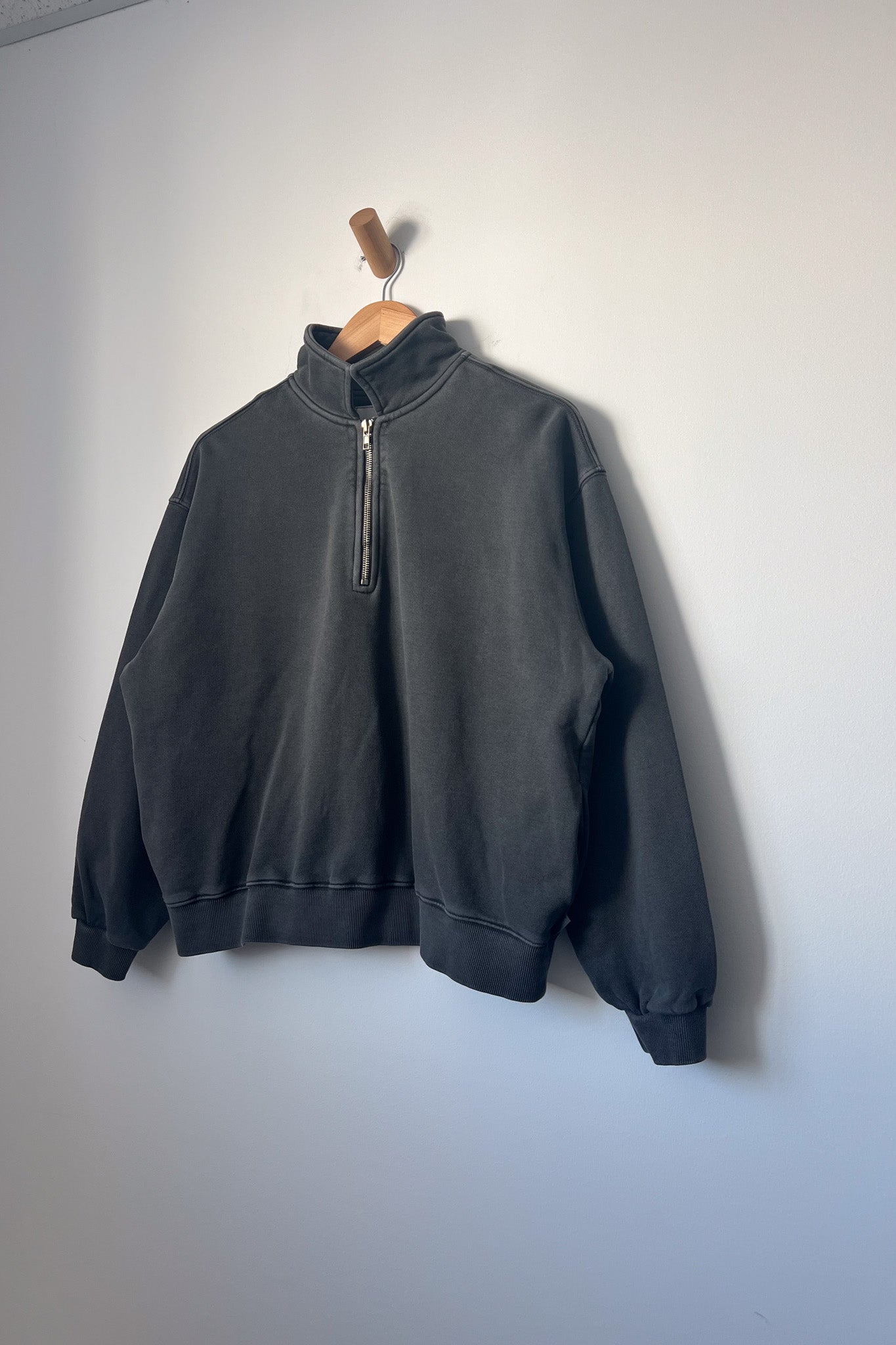 A charcoal-coloured quarter-zip sweatshirt hanging on a wooden hanger against a light grey wall. The sweatshirt has a relaxed fit, high collar, long sleeves, and ribbed cuffs and hem, with a visible side view.