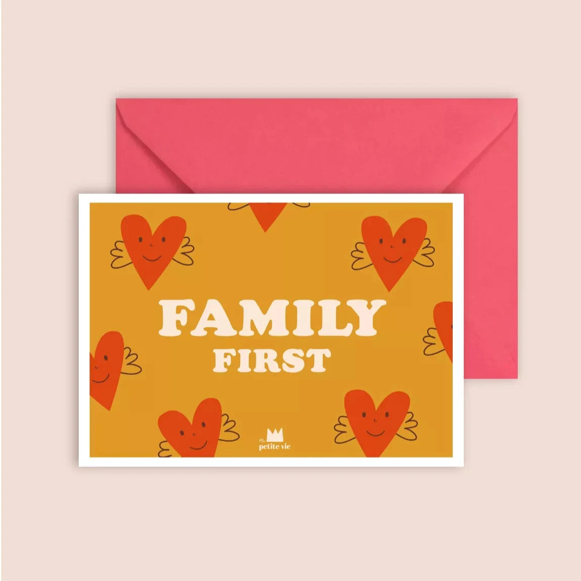 A postcard with a warm mustard-yellow background, featuring a pattern of illustrated red hearts with wings and smiling faces. The words "FAMILY FIRST" are printed in bold, white vintage-style lettering in the centre. The card is paired with a bright pink envelope.