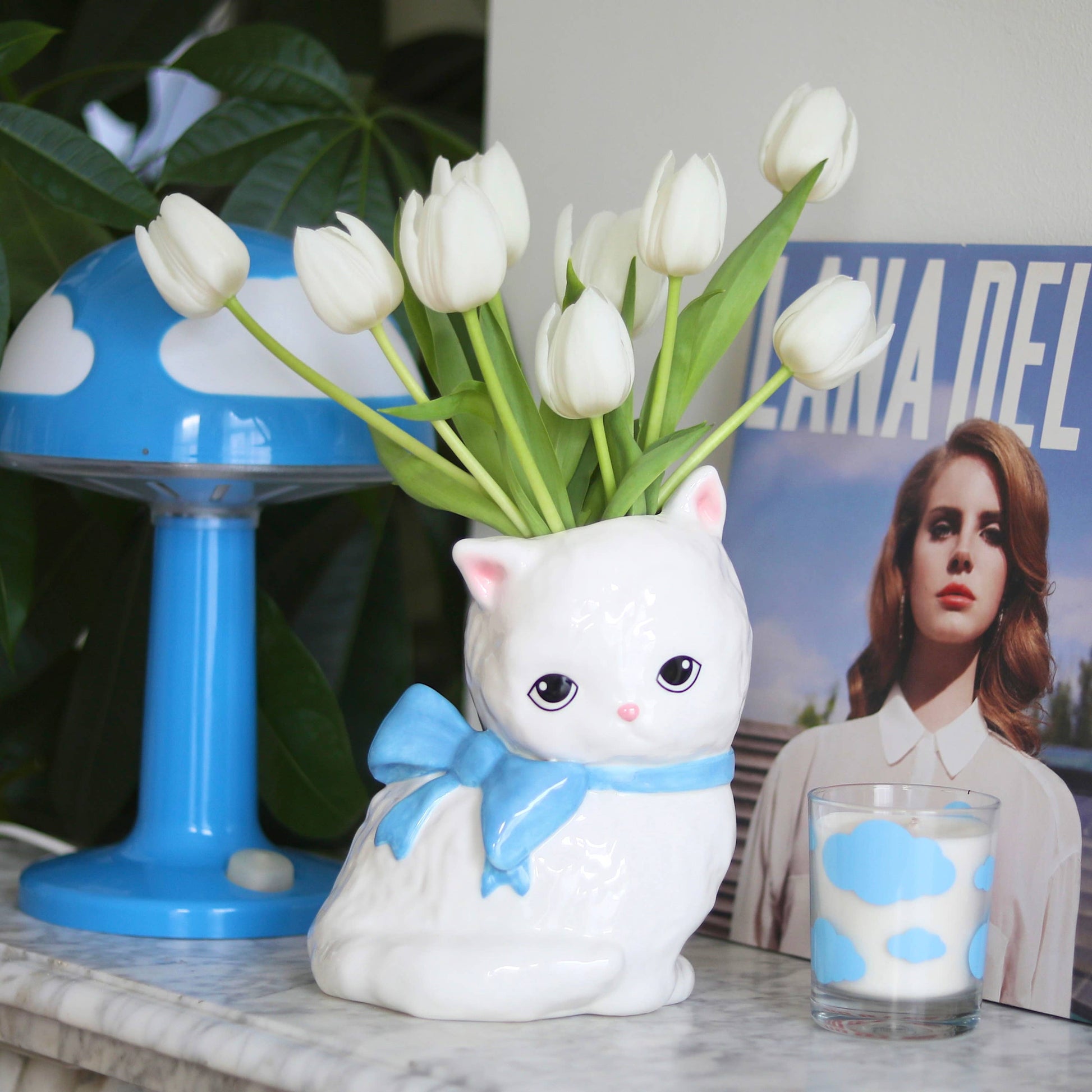 The white ceramic cat vase is placed on a marble surface, filled with fresh white tulips. It is styled alongside a blue mushroom lamp, a Lana Del Rey magazine cover, and a cloud-patterned candle.