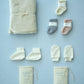 a flat lay on a baby blue backdrop. on the left is the fold waffle knit baby blanket, next to that is 3 pairs of socks, in pink, grey and cream. under the socks is the baby mittens and its draw string bag, then next to those is a pair of baby booties and its draw string bag