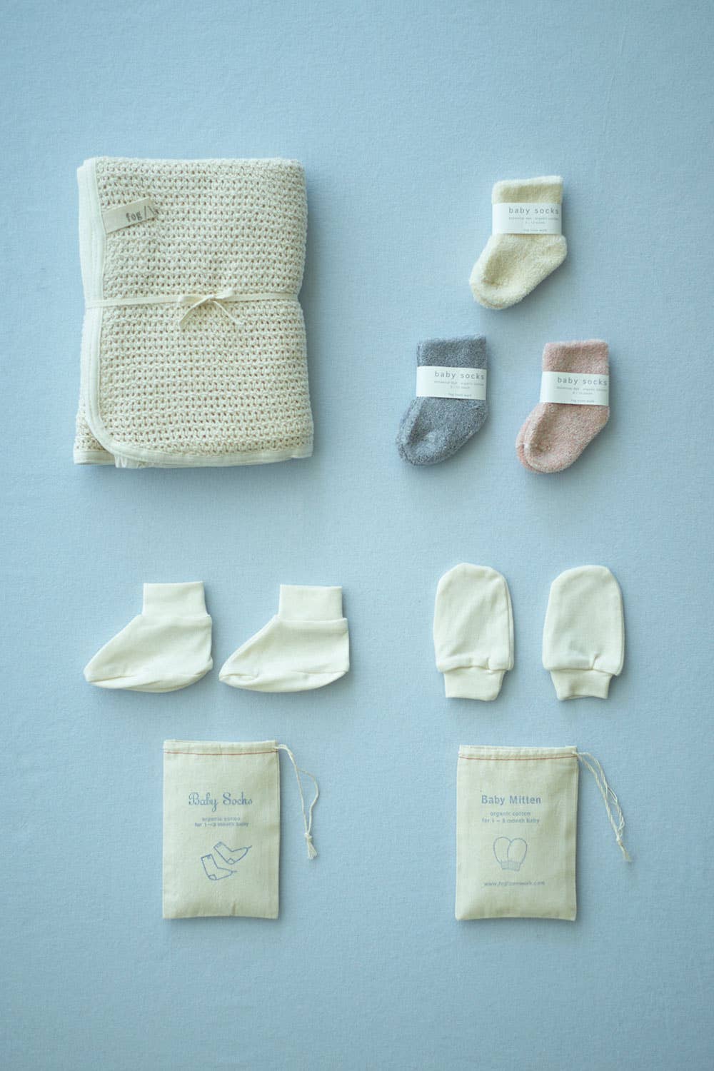 a flat lay on a baby blue backdrop. on the left is the fold waffle knit baby blanket, next to that is 3 pairs of socks, in pink, grey and cream. under the socks is the baby mittens and its draw string bag, then next to those is a pair of baby booties and its draw string bag