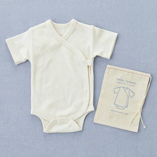 Cream cotton short sleeve wrap front baby romper, with poppers at the crotch and one at the cross over front. next to a draw string bag with an illustration of the romper and fog linen logo