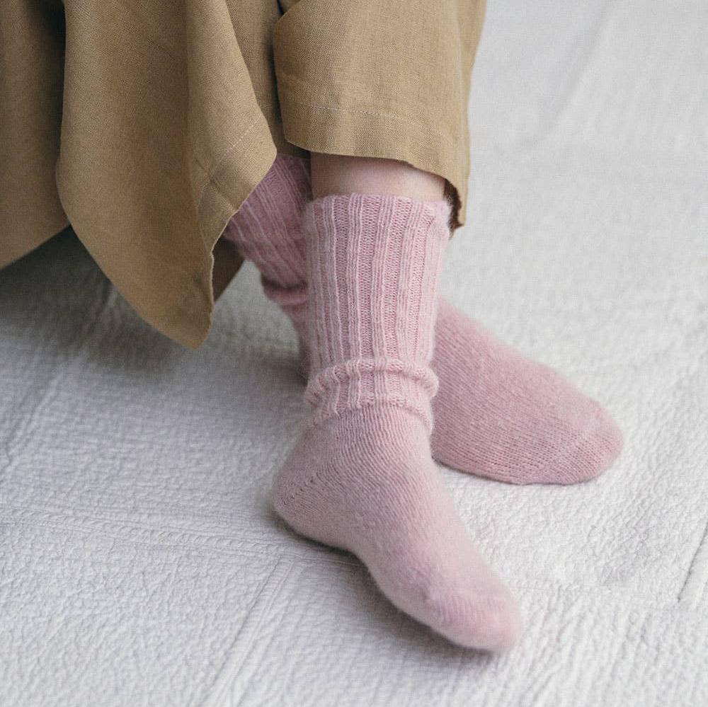 A woman ankles resting on a quilted white blanket, wearing a pair of baby pink mohair ankle socks with a chunky ribb pattern. her ankles are crossed and she is wearing a beige linen pair of wide culottes