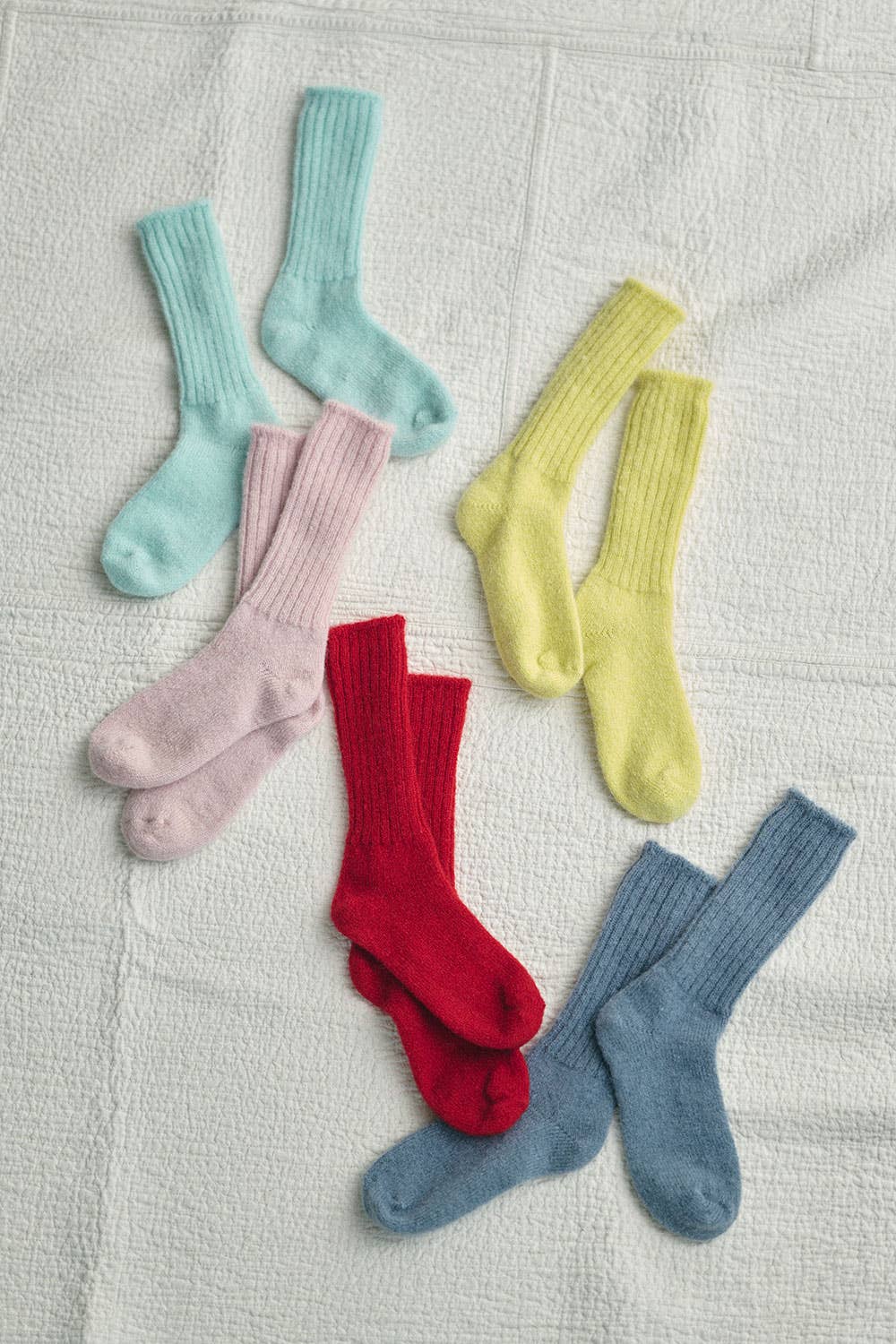 On a white quilted blanket, lay 5 pairs of Mohair socks scattered in a relaxed fashion on it. from top to bottom the colours of each pair are, light blue baby pink, yellow, red and denim blue