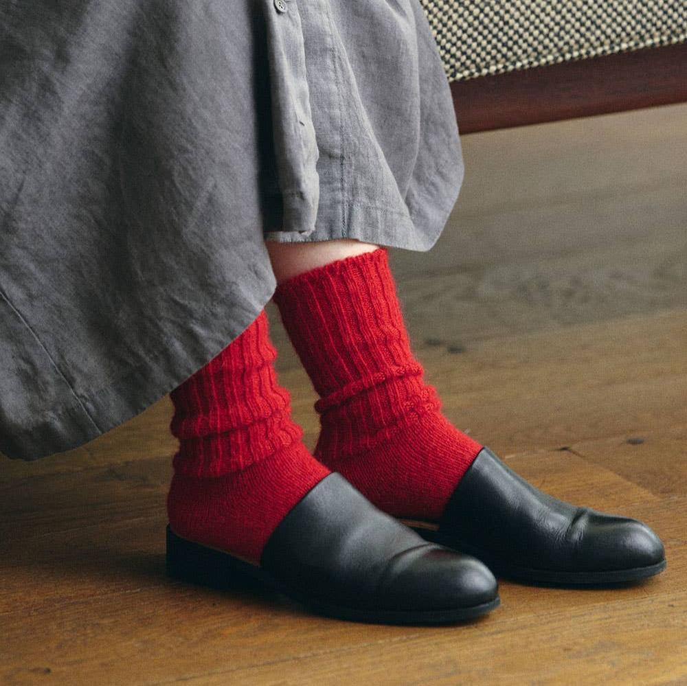 A woman wearing a pair of black leather low mules with a pair of red mohair socks with a chunk ribb texture. she is also wearing a grey linen dress. the background has old mid tone wooden floors