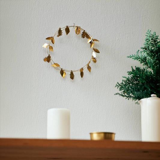 a delicate shiny brass garland with spread out leaves on a fine brass cord, wrapped in a double circle. displayed on a white wall and underneath is a wooden sheve with a candle and brass pot and a fern plant in white pot