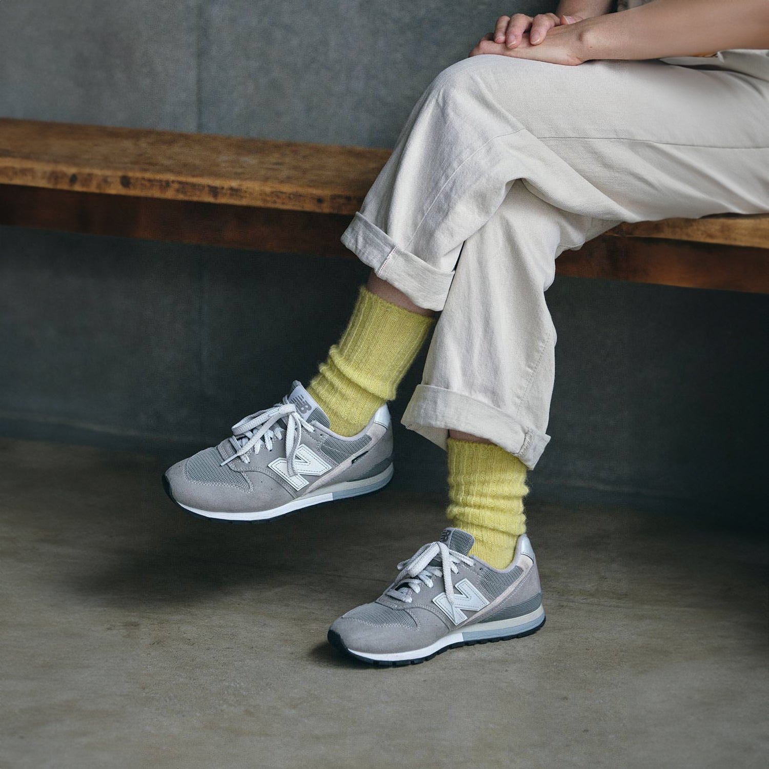 a woman sitting on a wooden bench with her legs crossed , wearing a pair of cuffed white denim trousers, yellow mohair ankle socks, and a grey pair of new balance shoes