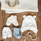 a flatly on a latticed wooden box. lays the two mittens, cream pair of socks and grey pair of socks, next to a tie Botton bear hat and above is the waffle baby blanket and a wooden baby tambourine