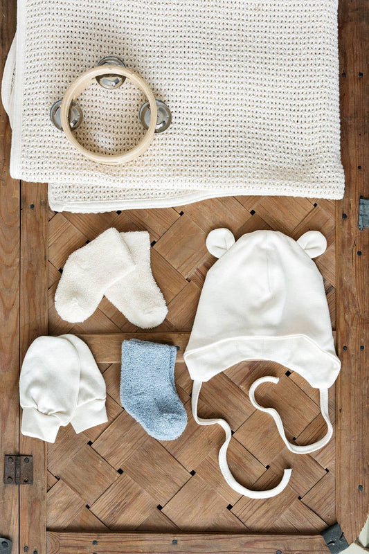 a flatly on a latticed wooden box. lays the two mittens, cream pair of socks and grey pair of socks, next to a tie Botton bear hat and above is the waffle baby blanket and a wooden baby tambourine