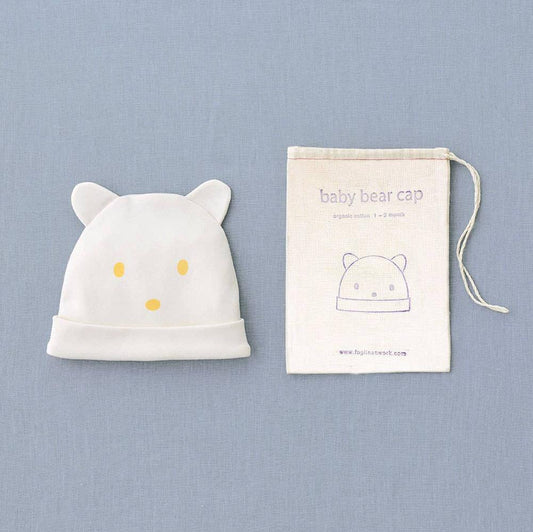 On a greyish blue backdrop, a baby white cotton hat with bear ears and yellow printed eyes and nose. Next to a little drawstring cotton bag with an illustration of the bear hat in it in blue lines with the fog linen logo